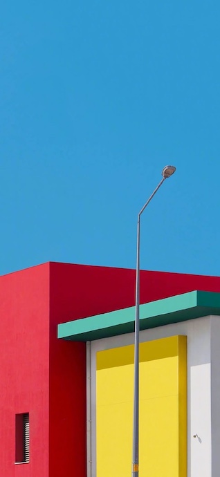 Minimalistische Architektur mit einer roten Wand, einem gelben Block und einem grünen Dachvorsprung vor blauem Himmel.