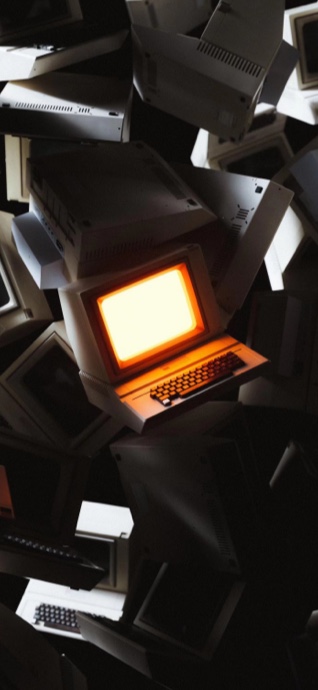 어두운 배경 속에서 따뜻한 주황색 빛을 내는 모니터와 쌓여있는 빈티지 CRT 컴퓨터들.