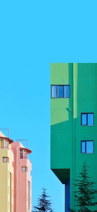 A vertical shot of minimalist green and pink buildings against a clear bright blue sky with a pine tree at the bottom.