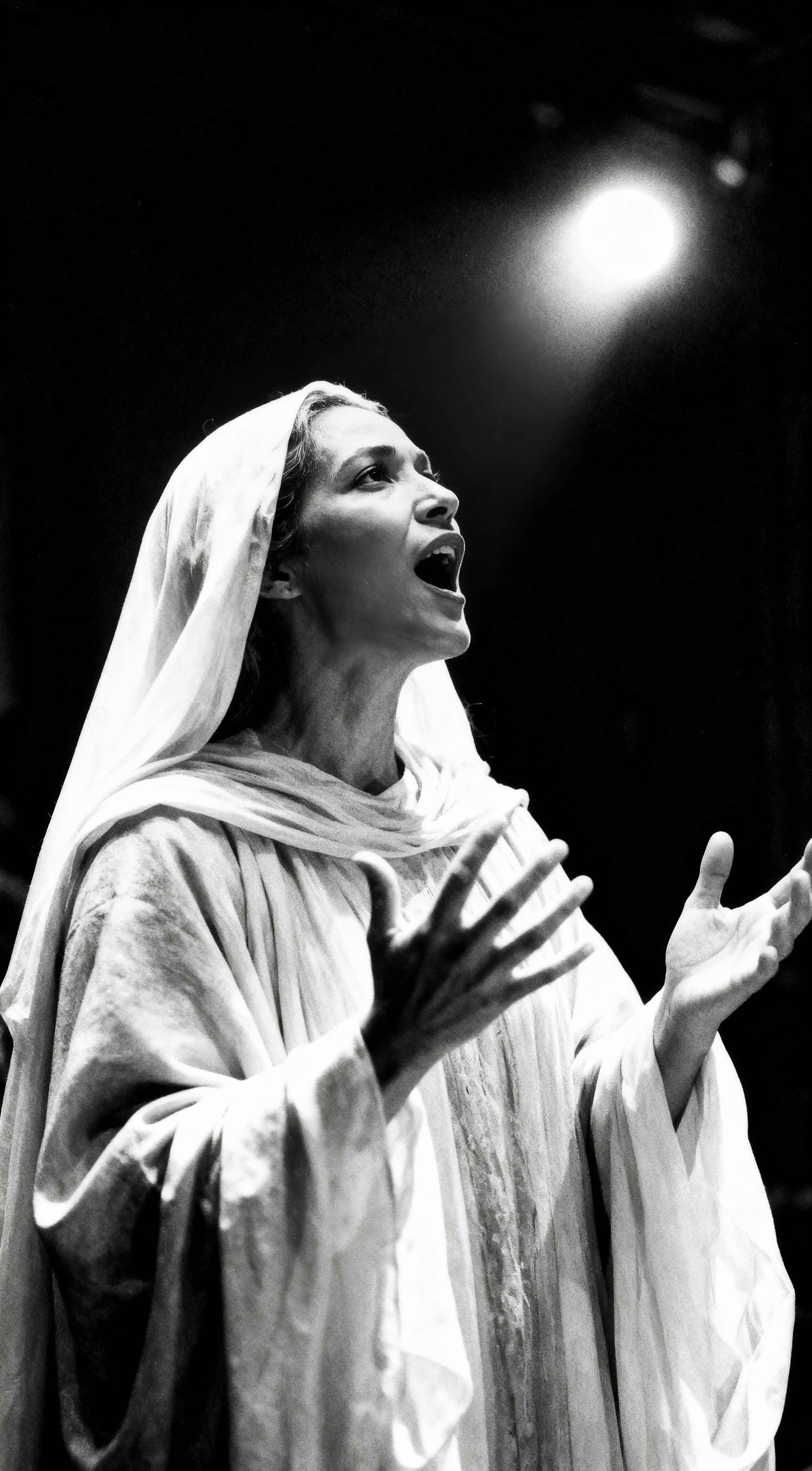 High-contrast black and white photo of Alexandria Ocasio-Cortez wearing a white veil, looking up and singing under a bright spotlight with hands raised in a theatrical pose.